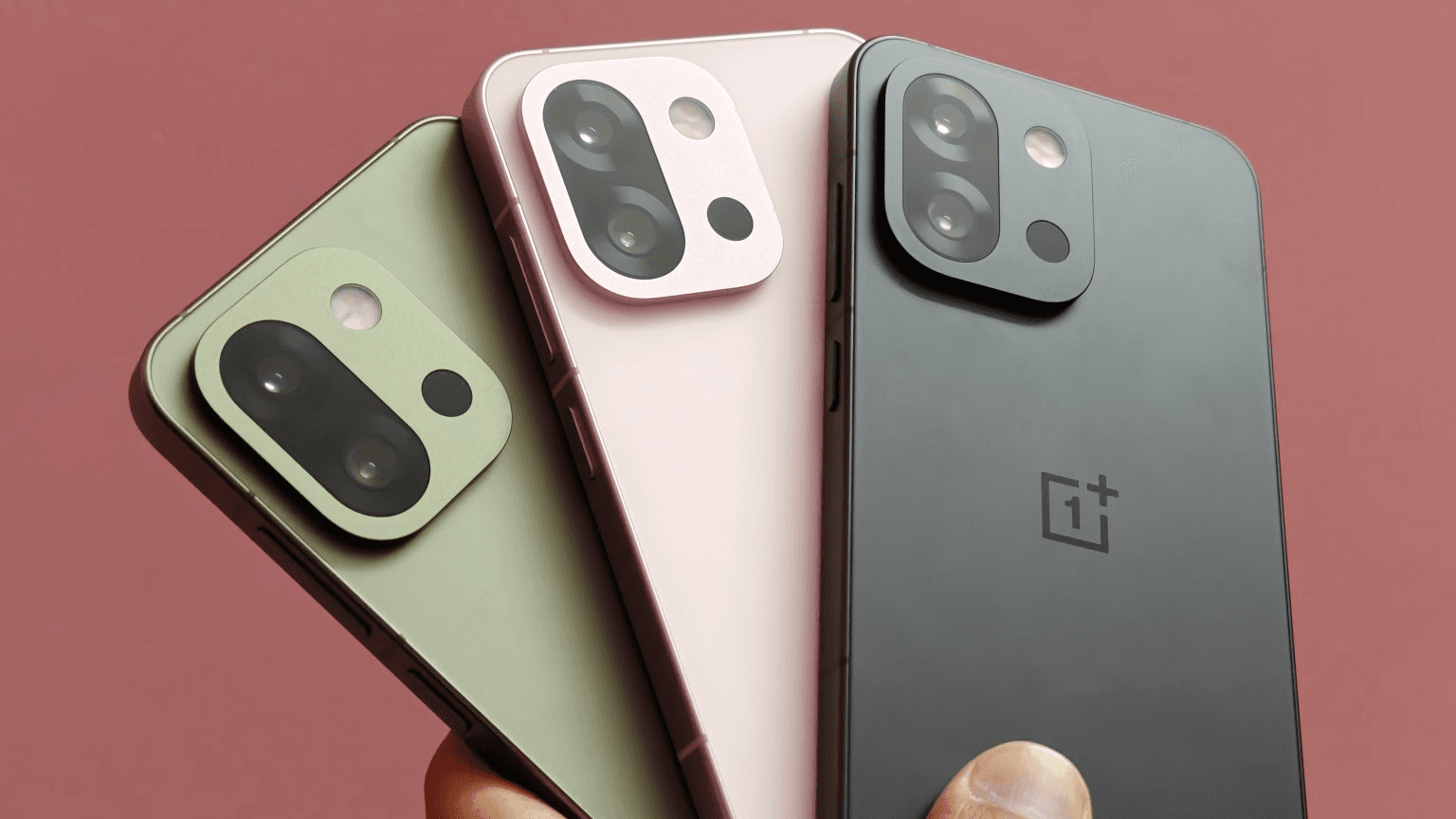 OnePlus 13s smartphones in black, pink, and green colors held in a hand against a pink background