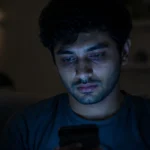 A tired person staring at a smartphone screen in a dark room representing Silent Scrolling Syndrome and Mobile Addiction.